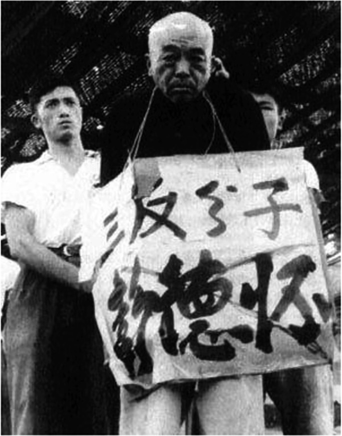 A photograph of an elderly man with a painted board around the neck that has text in a foreign language. 2 younger men hold the elderly man's hands behind his back.