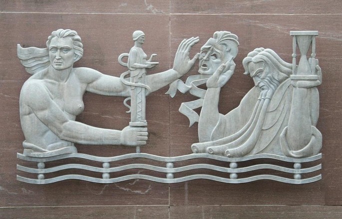 A sculpture of the Grim Reaper. On the right, is a man holding a sand clock in his one hand and a skull in the other. On the left is a man holding an arm out to stop the hand with the skull and a wand in his hand.