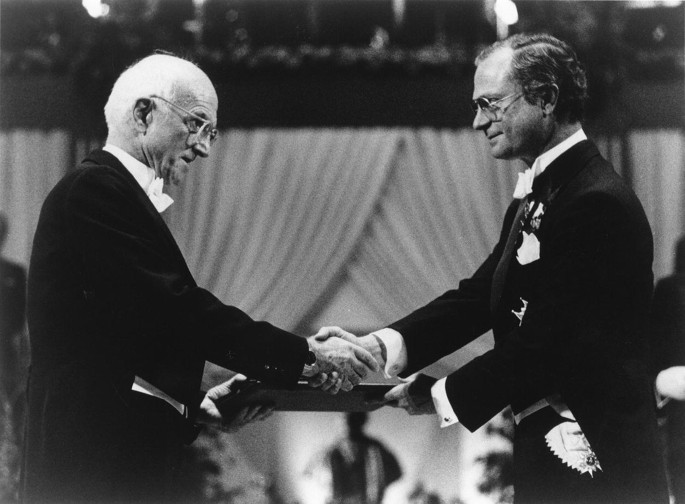 A photo of 2 men looking at each other and shaking hands. The men hold a document in their left hand.