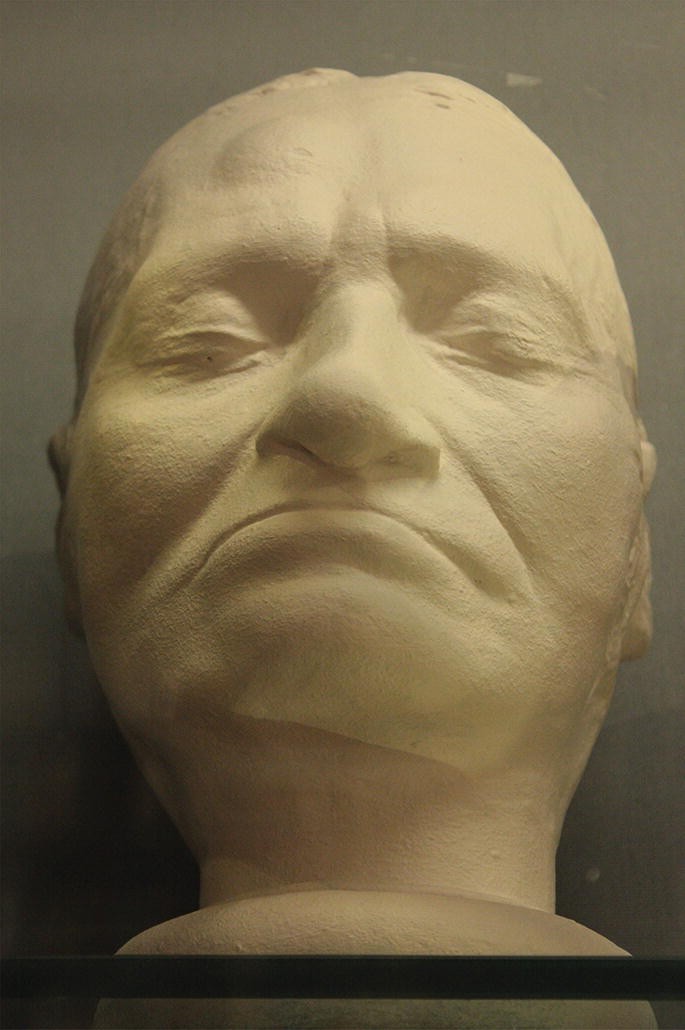 A photograph exhibits a close-up of a face mask of a person. The mask displays a bent nose, an uneven lower jaw, and smile lines.