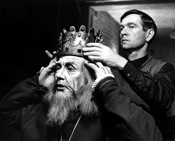 A photograph of a man stands behind an old man and places a crown over his head. The old man has a long beard and uses both hands to hold the crown over his head. Both men are looking at the left side.