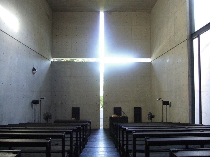 A photo of a Church room with a cross-opening on of the walls. Sunlight enters through the cross in the wall. There are benches arranged in rows in the room.
