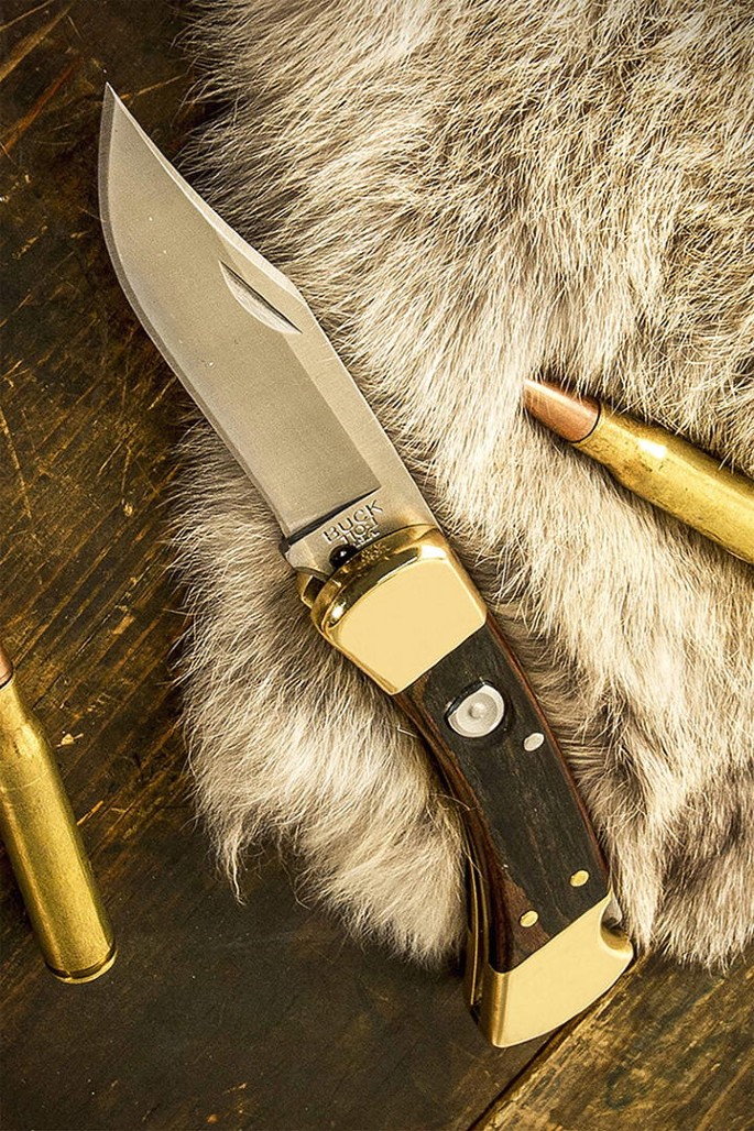 A close-up photo of a buck knife resting on a furry surface, surrounded by bullet casings.