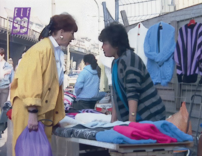 A photograph exhibits women interacting at a clothing stall in the market, with clothes displayed on the stall and hanging on the wall with hangers.