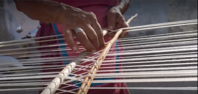 A screenshot from a film. It is a zoom-in of hands weaving threads on two wooden sticks.