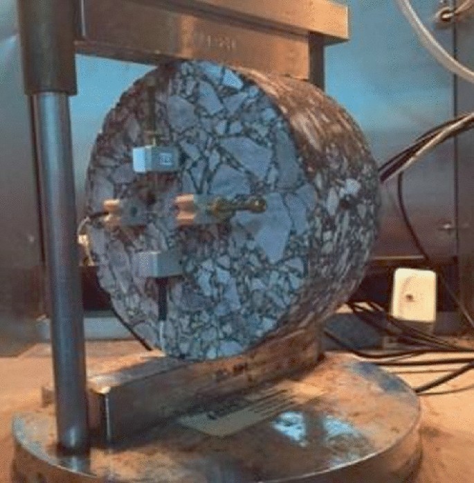 A close-up photograph of a tensile tester with a circular specimen attached in the center. The tester features a spherical base with a rectangular metal frame acting as a stand for the specimen.