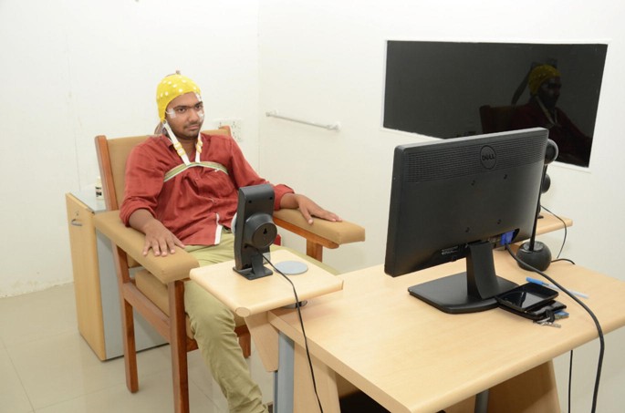 A man sits in a chair wearing B O S profiling gear on his head. There is a monitor and another device in front of him.