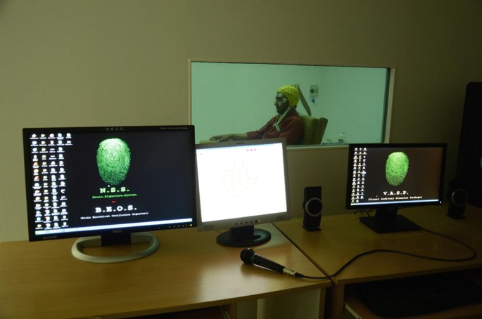 A set of 3 screens on a desk. The window on the wall behind the screens gives a view of the man who is the subject of the test. On the left screen is a wallpaper with a fingerprint, and below it are the acronyms N S S and B E O S. On the right one, the acronym is V A S P.