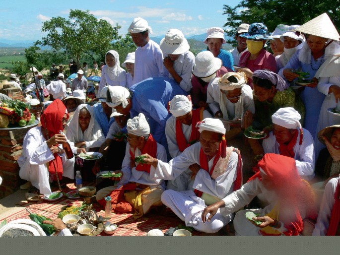 A photo of a few men in traditional attire with headdress performing a ritual outdoors. Many men and women surround the priests. Everyone wears a hat. Many utensils and other things are arranged on the ground.