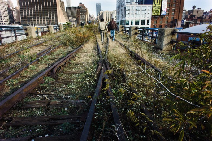 A photograph of the High Line rail tracks. The tracks are covered with bushes. The sleepers are damaged, and the fixtures are corroded. Two people walk along the tracks.