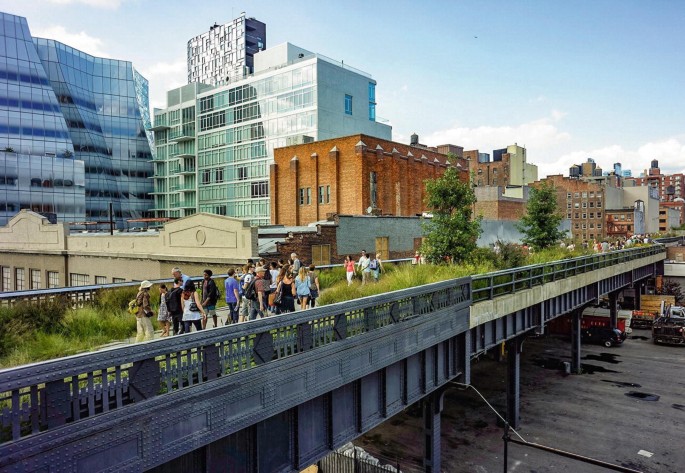 A photograph of a renovated High Line rail tracks. The tracks are covered with bushes. A group of people walk along the tracks. An old building and a few newly constructed buildings are in the background.