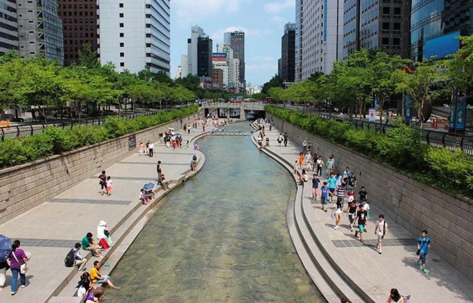 A photograph of Cheonggyecheon Stream with areas for sitting and walking on both sides of the river. People walk and sit in the areas.