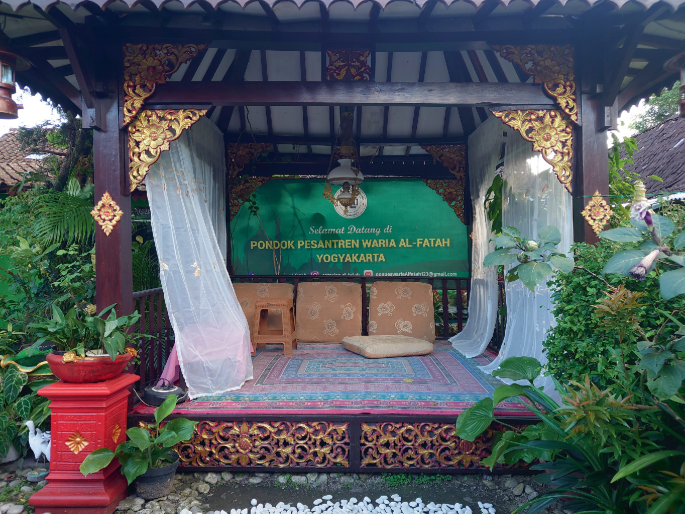 A photograph of a gazebo with a mattress, cushions, and stool inside. The board in the background exhibits text in a foreign language. The surrounding has plants and flowers.
