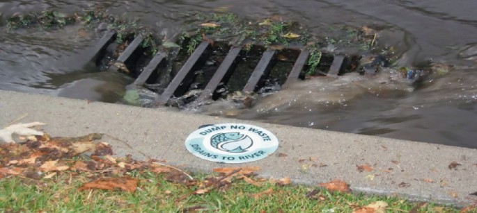 A photograph of a storm drain. The stormwater rushes into the drain. The drain has a metal cover with openings. A sticker on the side of the pavement has a logo with text that reads, dump no waste, drains to river.