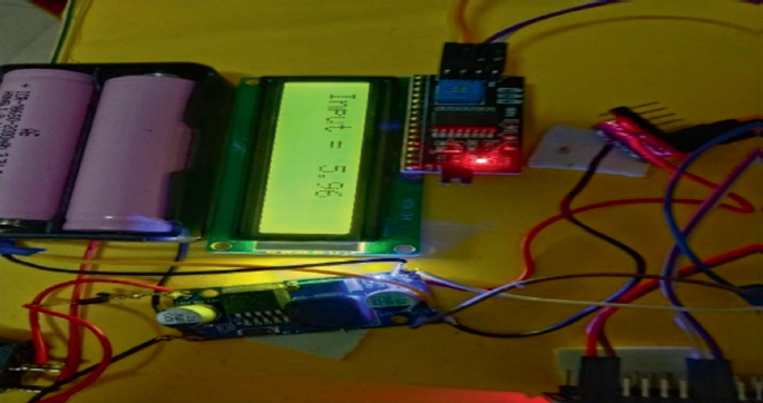 A photo of a voltage output from the charged battery has a display screen with data that reads, input equals 5.96 and voltage booster, batteries, 12 C L C D, and other electronic devices connected with wires.