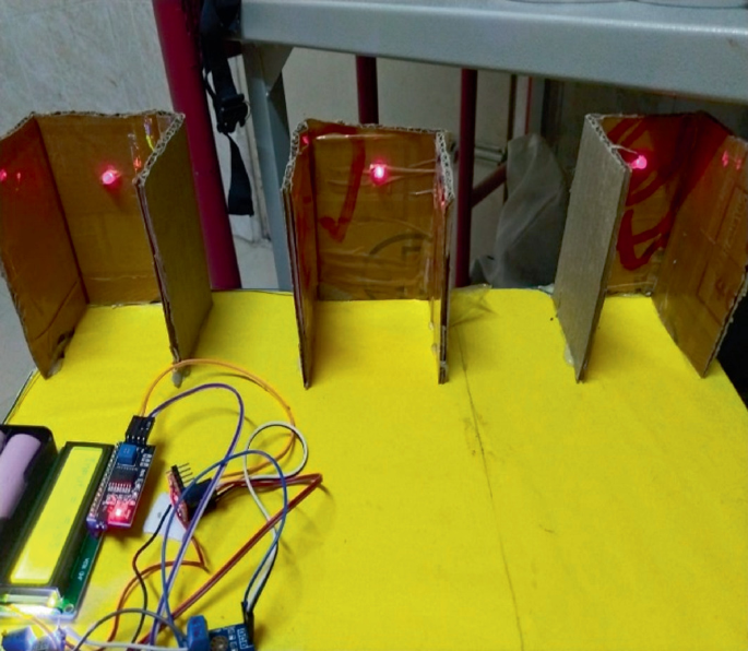 A photo of a model based on the piezoelectric effect has 3 cardboard boxes open from 3 sides top, bottom, and front, vertically placed in a line side by side with glowing L E D bulbs one in each, a voltage booster, I2 C converter for L C D display connected with wires.