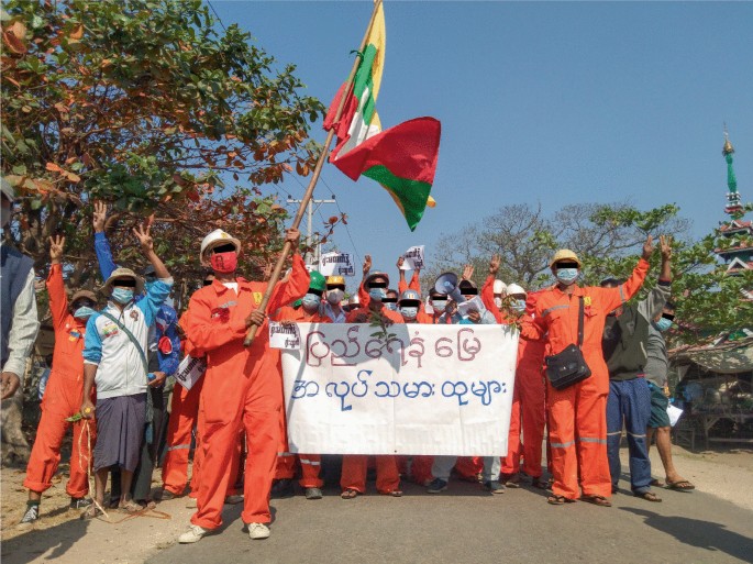 A photograph of uniformed union members. They hoist the flag of Myanmar and carry banners. The text is in a foreign language.