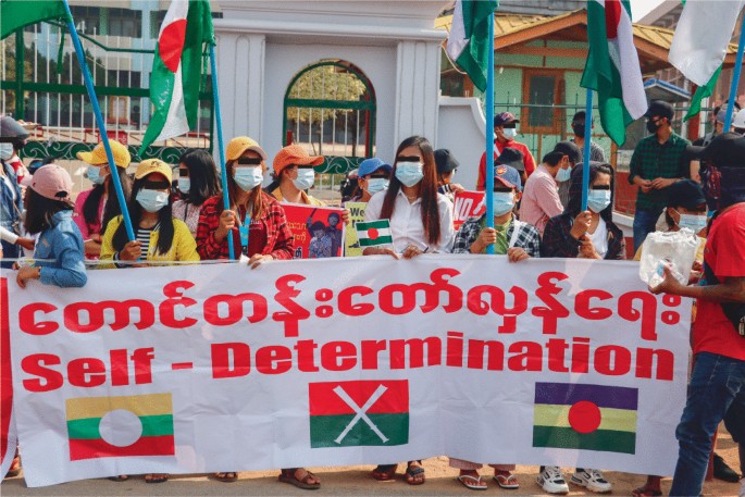 A photograph of a group of people hoisting the flags of Myanmar. They hold a large banner with text that reads self-determination.