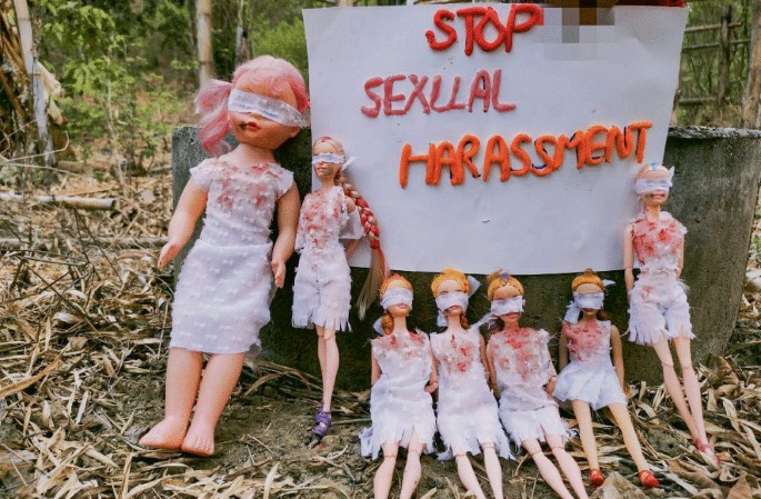 A photograph of dolls dressed in white with spots of blood, blindfolded, and tied arms. There is a poster that reads stop sexual harassment.