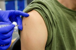 Hands wearing blue gloves insert a vaccine needle into the arm of a person wearing a green shirt rolled up to their shoulder.