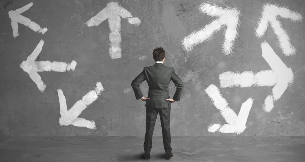 Mann steht vor grauer Wand, auf der verschiedene Richtungspfeile abgebildet sind