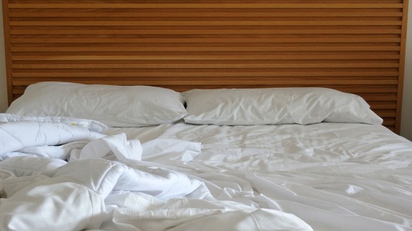 An unmade bed with qwhite sheets against a light brown headboard. 