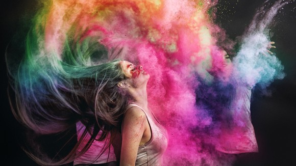 A white woman wearing a white tank top is photographed while splashing her hair back, spreading colourful holi powder around her. The picture is taken from a side angle, showing the woman’s profile. The background is black, and the holi powder is blue, green, orange, and pink.