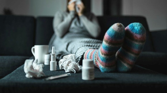 Woman sneezing and blowing nose with tissue and handkerchief. Sick and ill person with flu, cold medicine and woolen socks. Fever, virus or infection concept. Sitting on couch at home in winter.