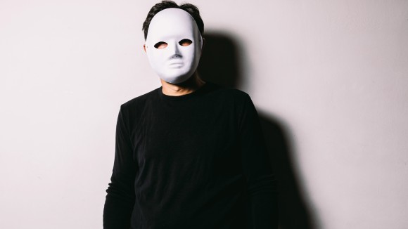 A person wearing a black jumper is pictured from the waist up, and stands in front of a white wall with arms at their sides. They are wearing a white mask covering their entire face and short hair can be seen at the top of their head.  
