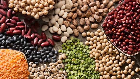 Up close image of several varieties of legumes (chickpeas, lentils, beans, peas) in bowls and on a table.