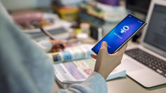 Close up view of a teenage girl interacting with a multimodal AI chatbot on her smartphone while studying at a desk with a laptop and notebook. 
