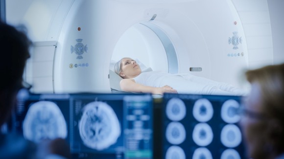 Woman laying in MRI machine with blurred computer screens with brain scans and two people on the side.