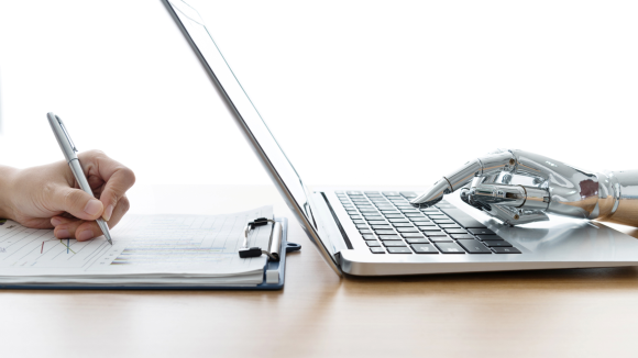 On the right hand side of the image, there is a side view of robot hand typing on a laptop; on the left hand side of the image there is the image of a human hand writing on paper using a pen