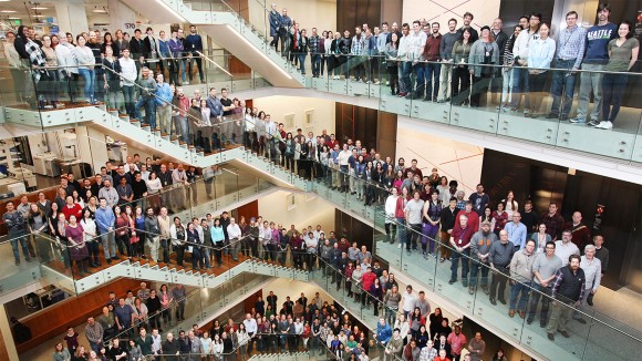 A crowd of people are gathered on the steps in the atrium of a lab, smiling up at the camera.