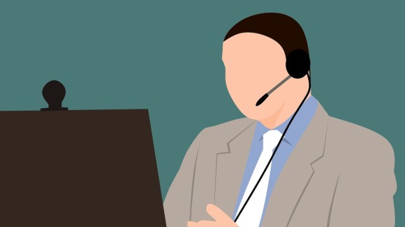 Cartoon drawing of a light skinned person with short hair, a grey suit, blue shirt, and white tie sitting in front of a computer monitor with a webcam. They wear a headset and are styled to look as though they are on a video call.