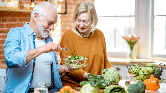 An elderly man and woman happily prepare healthy foods in a clean kitchen.