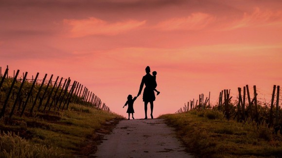 Silhouette of mother walking with two young children along path in an orchard field at sunset.