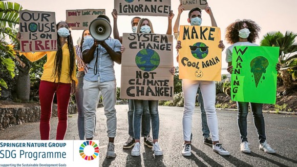 Seven young people of diverse ethnicities stand in the middle of a road holding protest-style signs reading messages like “Our house is on fire!!”, “Evidence over ignorance”, “System change, not climate change”, “make earth cool again”, “there is no planet B”