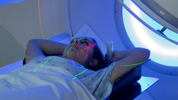 Young woman in hospital gown lying down on scanner with green and pink lights visible on her arms and face.