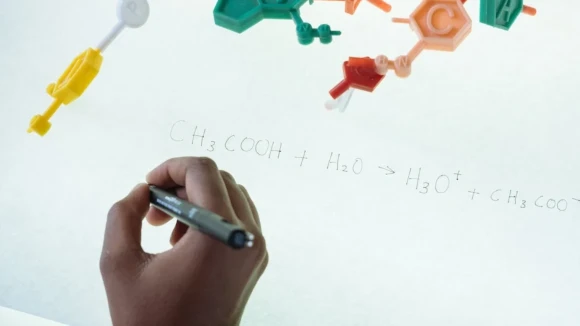 A chemical formula is written down on a sheet of paper, with a hand holding a pen visible in the lower corner of the image. A plastic model of a chemical molecule is visible in the top left corner of the image.