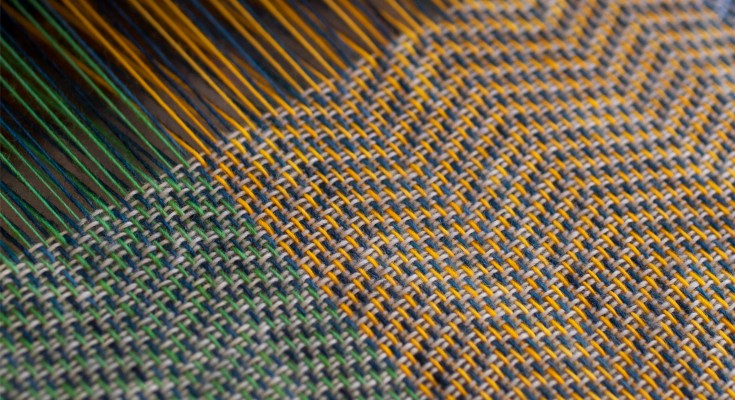 A loom on which cloth is woven from multicolored threads. The roman numeral V emerges as separate threads come together to form a patterned fabric.