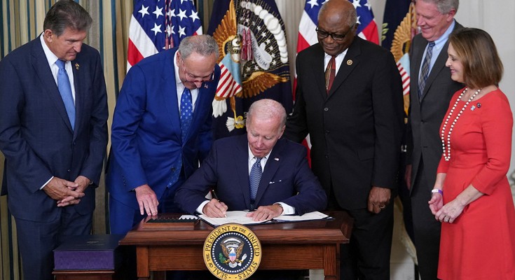 President Biden sits at a table with the US presidential seal on it, signing a document