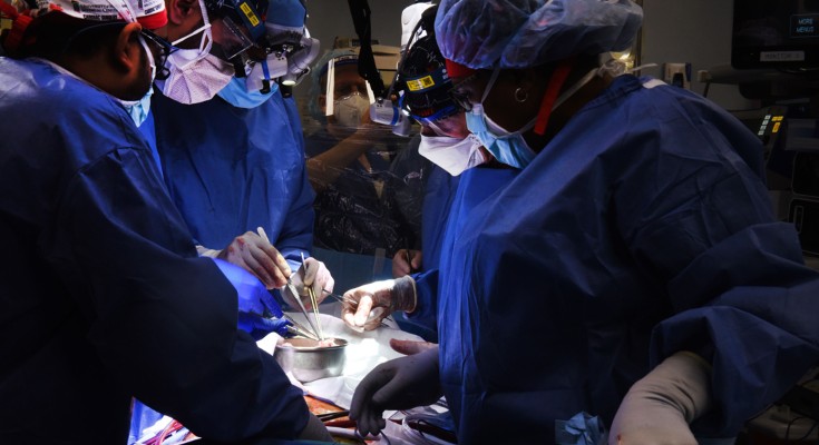 A surgical team of five people stands over a patient, with medical instruments in the foreground
