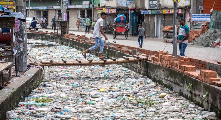 Man walks over a small makeshift bridge across a canal which is blocked by piles of plastic waste