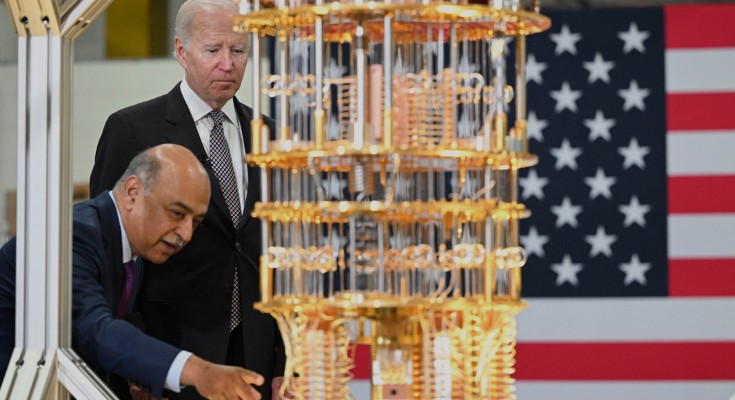 President Biden stands examining a bright metal quantum computer with IBM CEO Arvind Krishna, who is leaning over to point something out. There is a US flag behind them
