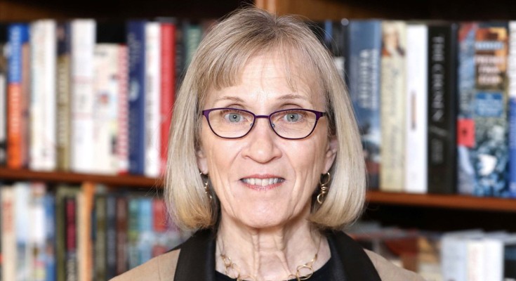 Claudia Goldin, a woman with bobbed light hair and glasses, looks to camera and stands in front of a bookcase