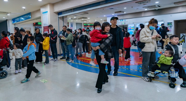 A crowded hospital in China with many parents and small children. In the foreground, a worried-looking woman holds a small child