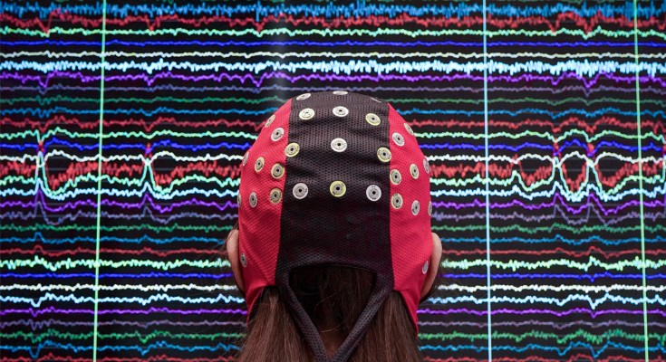 A person wearing a brain-computer interface cap looks towards a screen with graphical lines in many colours