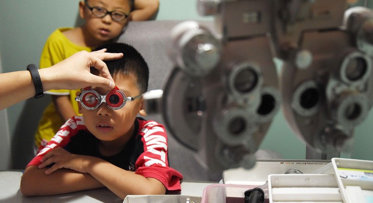 A child in the foreground wears eyesight test glasses, while an adult hand replaces the lens. Another child sits in the background