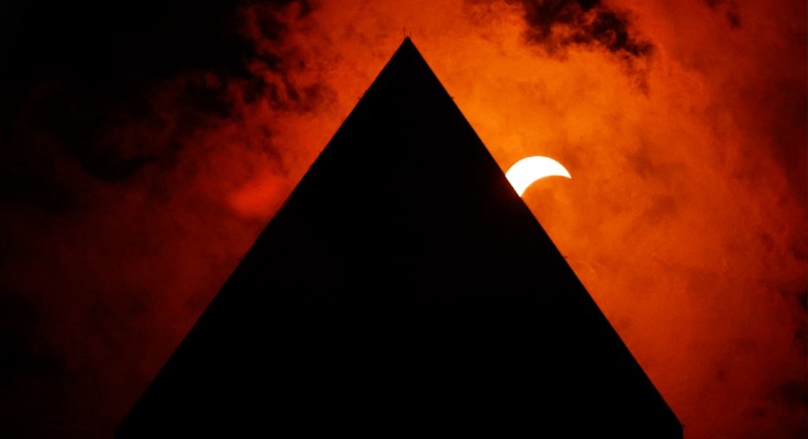 An eclipse behind a monument. A crescent moon pokes out from behind the monument, which appears surrounded by red smoke